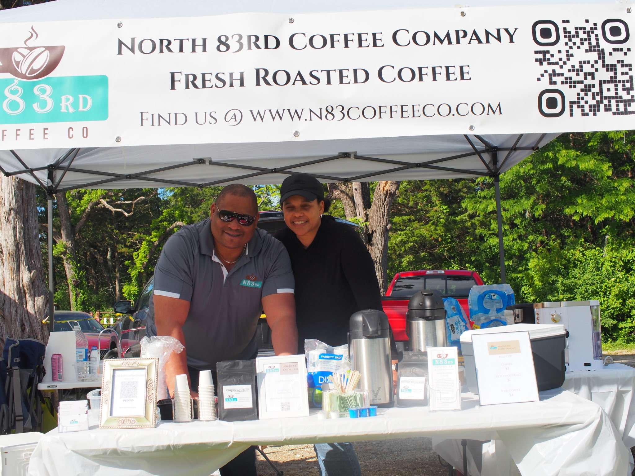 Two people at a North 83rd Coffee Company outdoor market stall, with coffee products on a table under a company banner
